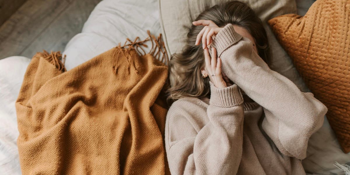 Woman resting on bed A white woman with brown hair and her face obscured by her hands lays face up on her bed, flanked by orange pillows and blankets.