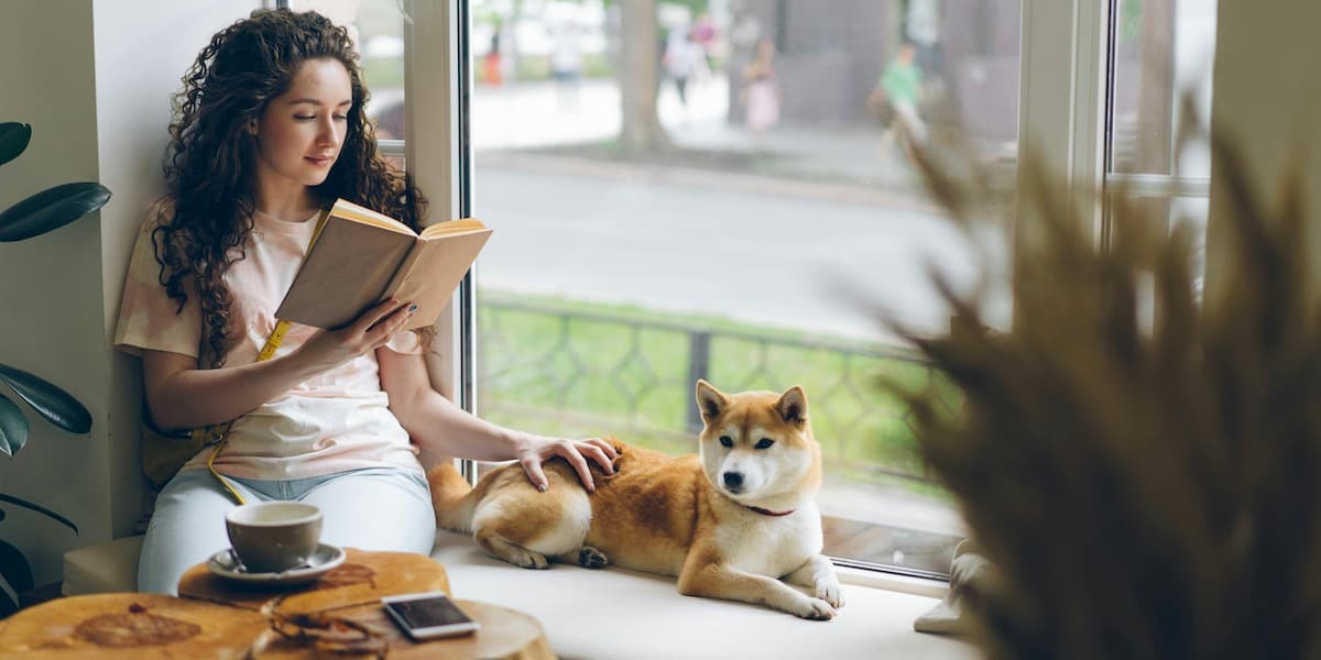 Woman reading at window A white woman with long dark curly hair sits in the window of a coffee shop reading a book and petting her shiba inu dog, who lays in the window seat beside her.