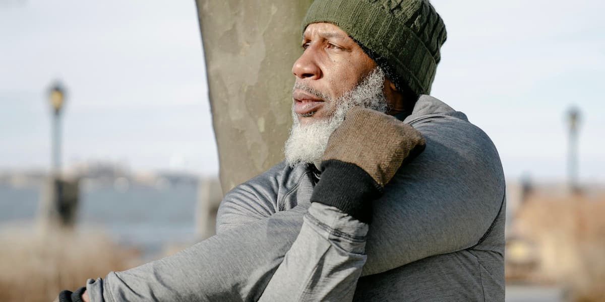 Man stretching pre-workout An older Black gentleman wearing a knit cap, long sleeve shirt and gloves looks away from the camera and stretches as he warms up to work out in a New York City park.
