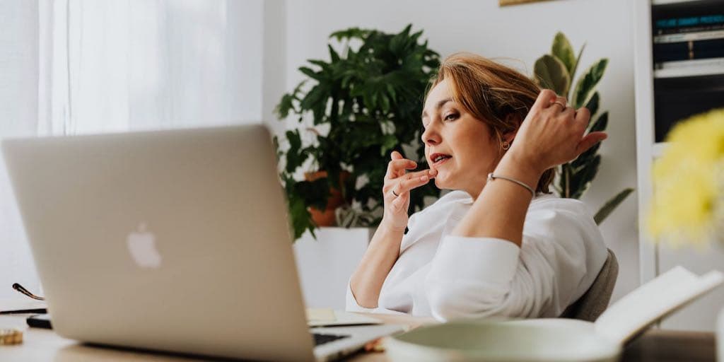 A young white woman with short red hair wearing a white shirt sits at a table in a bright white room in front of a computer and touches her lip as she gazes into the screen.