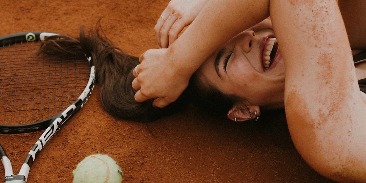 Woman laughing on tennis court A young white woman with brown hair lays covered in dark red tennis court clay as she lays on the court and laughs, a racket and balls nearby.
