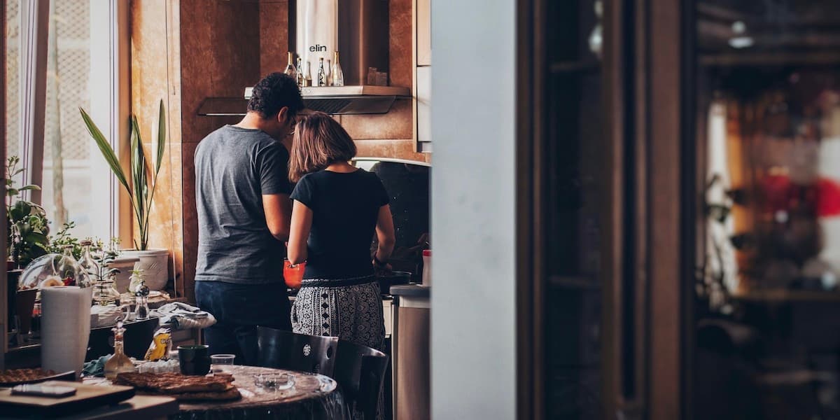 A white couple stand together at their stove in a warm, cluttered kitchen with many wood accents and comfortable clutter.