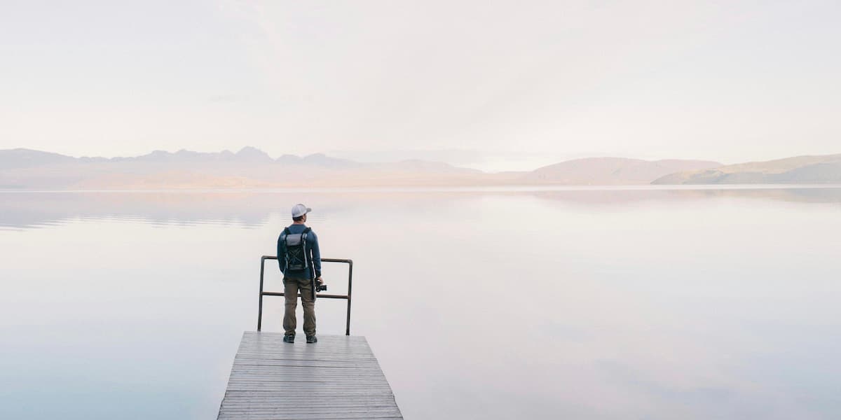Man on dock A pastel photo shows a man wearing hiking clothes from behind. He's standing on a dock in the distance, looking out over the foggy water.