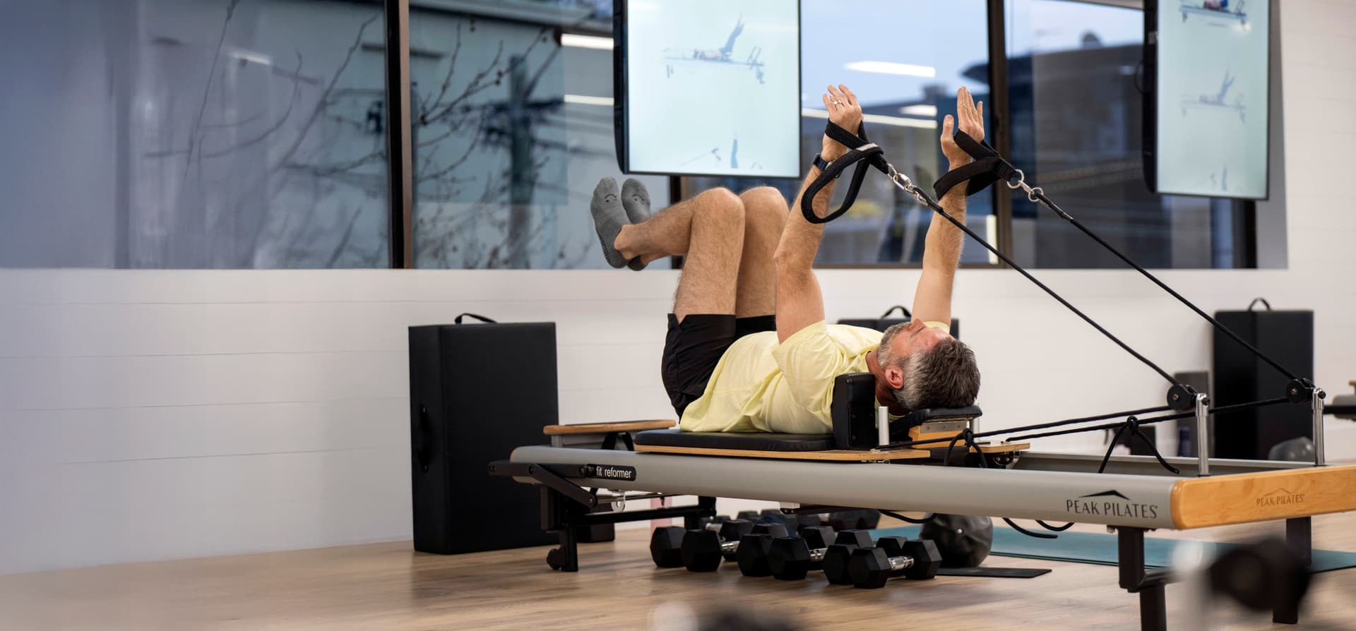 A man exercises on a Pilates reformer, using straps for his hands and feet, in a bright, modern studio.