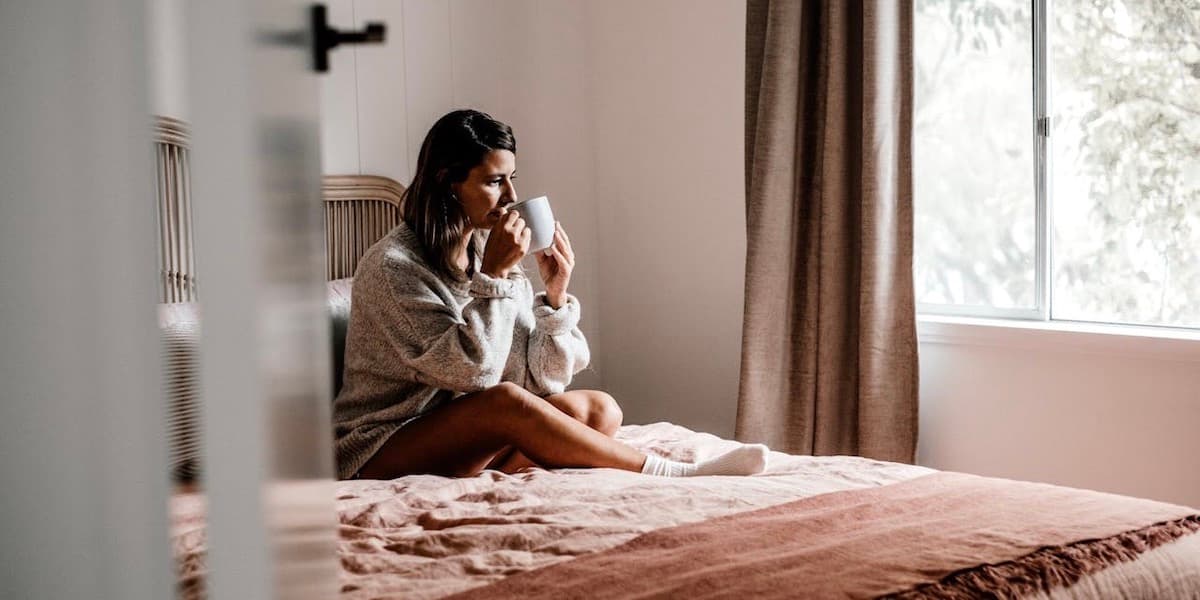 A young white woman with long dark brown hair sits on her bed wearing a sweatshirt and shorts, sipping from a mug.