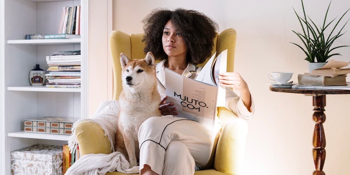 Woman reading in chair with dog An attractive young Black woman with an Afro wearing button down pajamas sits in a gold wingback chair next to a bookcase with her dog, holding a paper, both of them looking at the same point off camera.