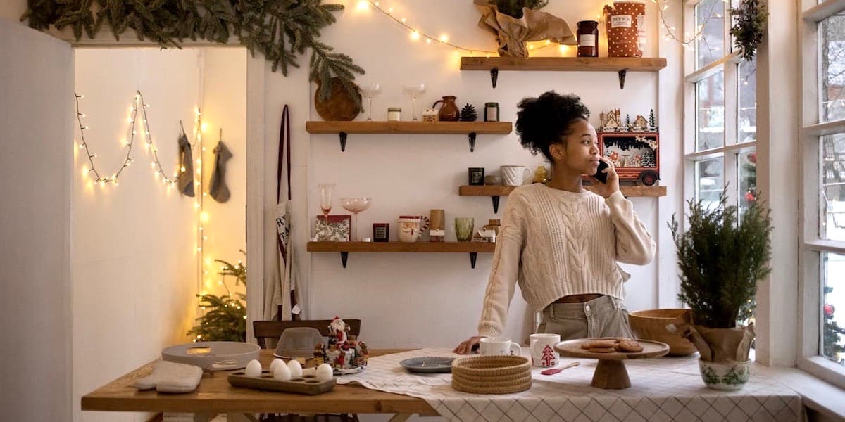 A young Black woman wearing a sweater talks on her phone while looking out her kitchen window in a cozy kitchen decorated with twinkle lights and pine boughs and small tress. Open pine shelves are behind her and the counter in front of her is spread with baking materials.