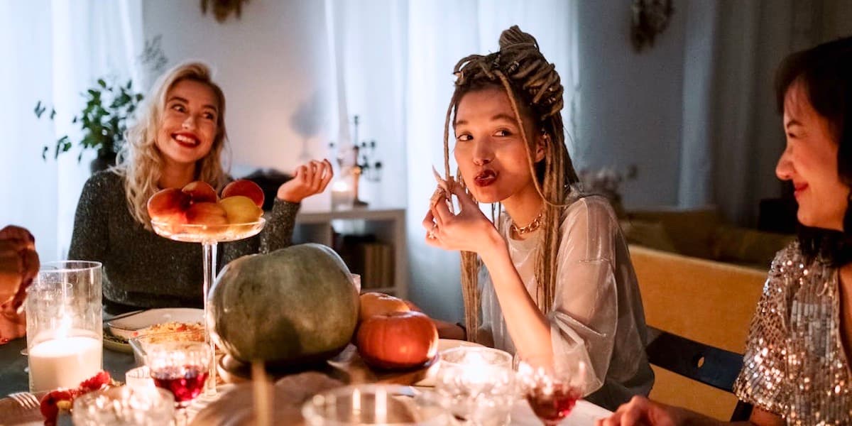 Friends at holiday dinner party Young stylish friends gather at a dining room table for a holiday party, smiling and eating with food and wine glasses scattered on the table, under twinkling candle light.