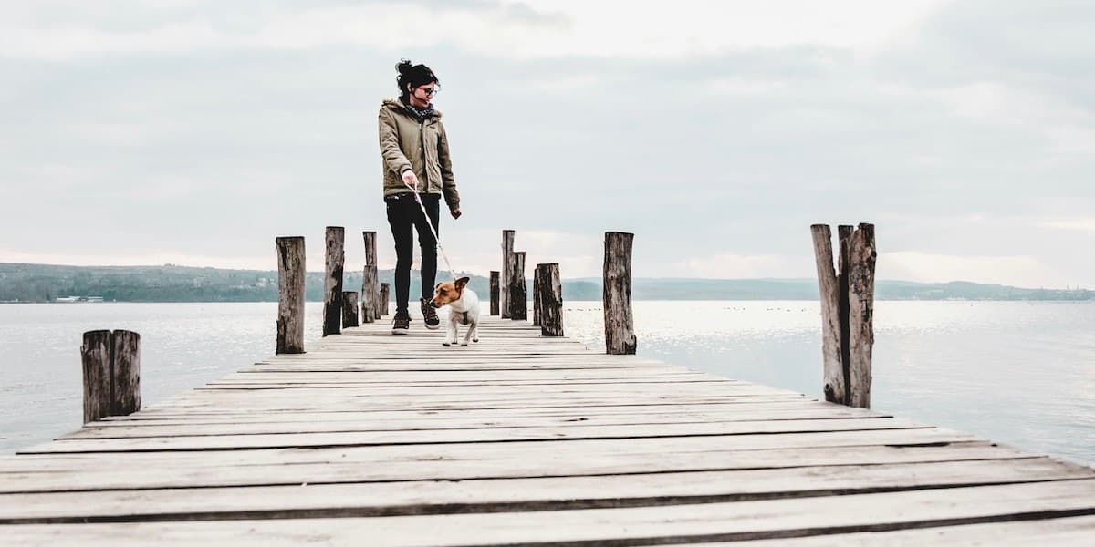 Woman and dog on dock A young white woman in winter clothes stands at the edge of a dock with her small terrier dog, a wide expanse of water behind them, walking back toward land and the camera.