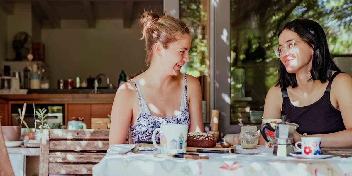 Two young women sit at an outdoor cafe table, drinking espresso and eating pastry, smiling as they chat.