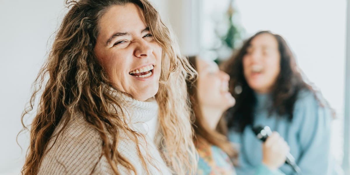 A woman with long wavy blond hair wearing a warm turtleneck laughs into the camera as her friends belt out karaoke into a microphone, blurred in the background behind her.