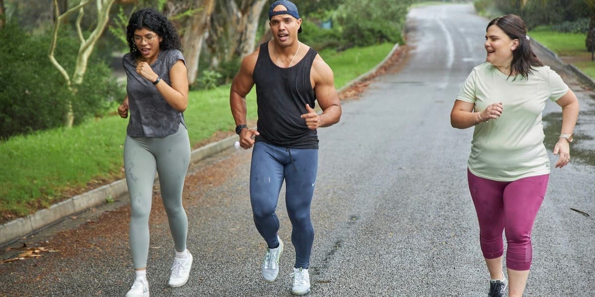 Three young people of various body sizes in workout clothes run together on a paved road with grass and trees on the side.