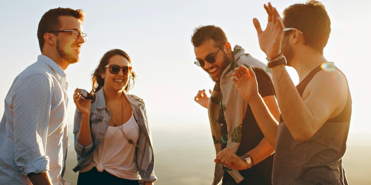 Four young adult friends in summer clothing smile and laugh on the beach during sunset, with the light creating spots and shadows around them.