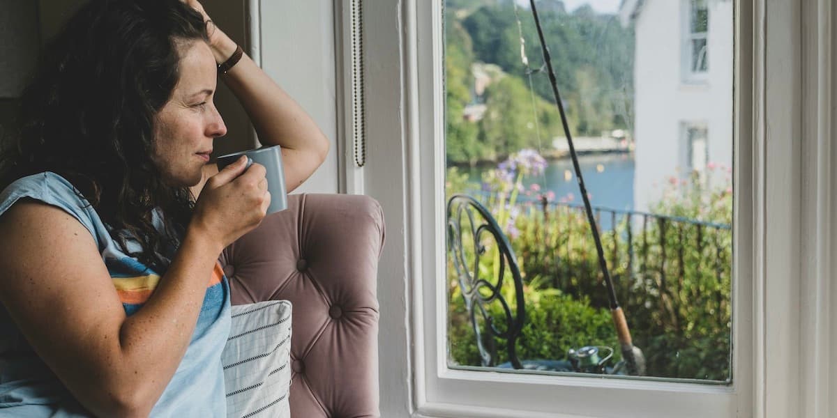 A woman wearing a colorful t-shirt sits in a window seat at home, holding a mug to her lips and staring contemplatively out the window, where water, greenery and a fishing rod leaning against a chair can be seen.