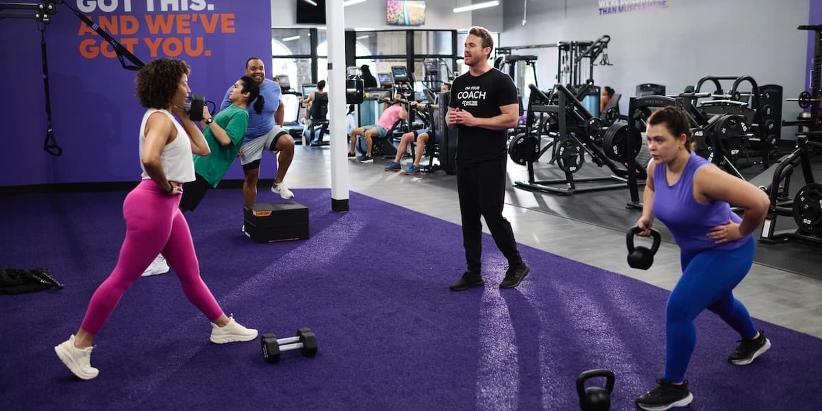 Two women lift weights with the help of a trainer at Anytime Fitness while other people exercise in the background. Photo by Anytime Fitness.
