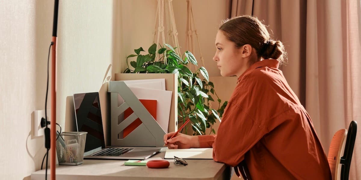 A young woman in a large red shirt with her hair tied back sits at a desk and writes on paper while looking into a computer screen.