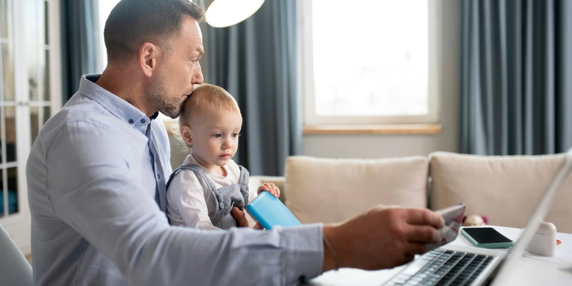 A young father in a button-down shirt kissed the head of the infant child in his lap, while both sit at a table in a living room in front of a computer, while he holds a credit card toward the screen with his free hand.