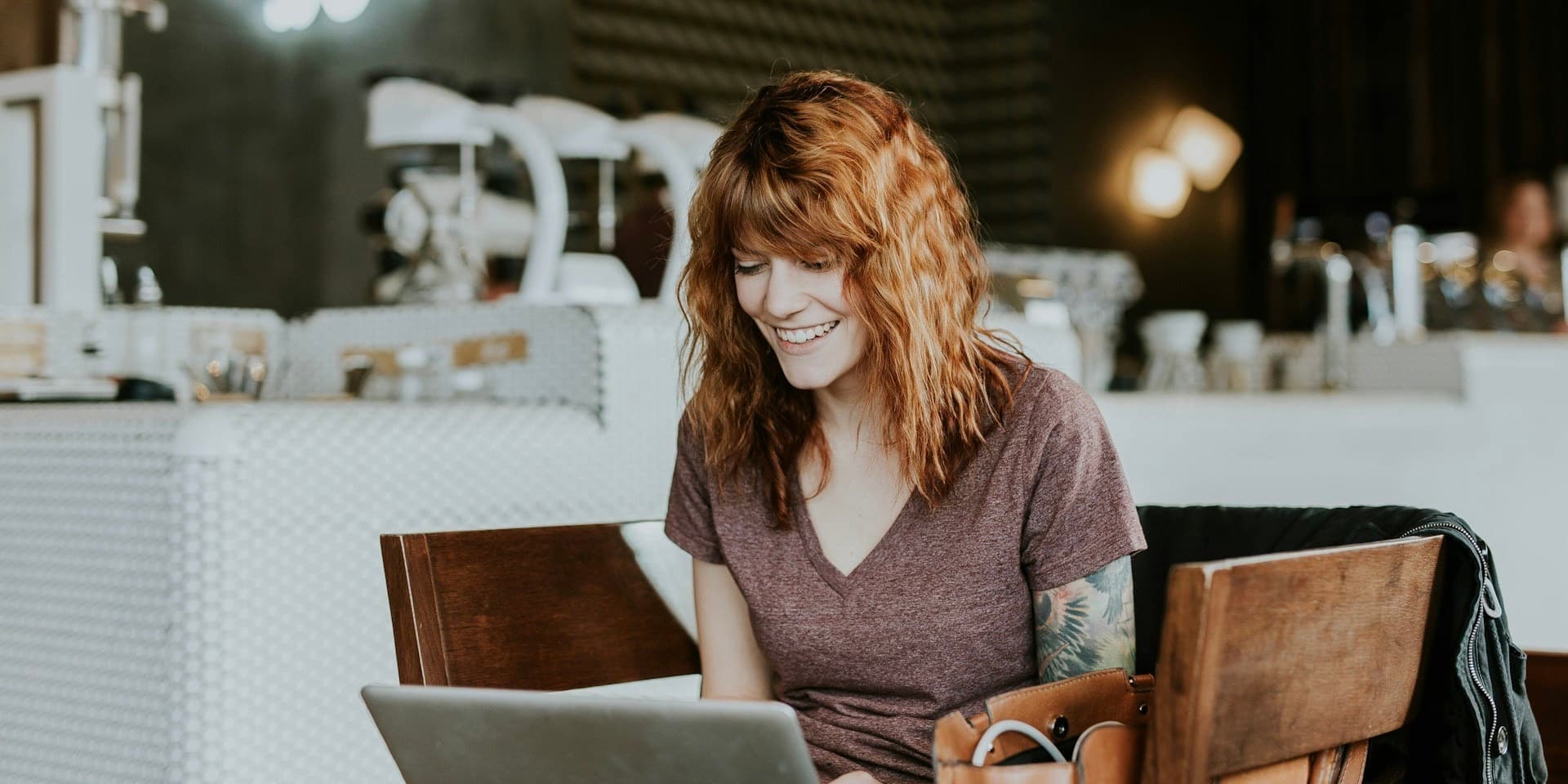 A young woman with wavy red hair and tattoos wearing t-shirt sits in a trendy coffee shop, smiling as she looks down into a laptop.