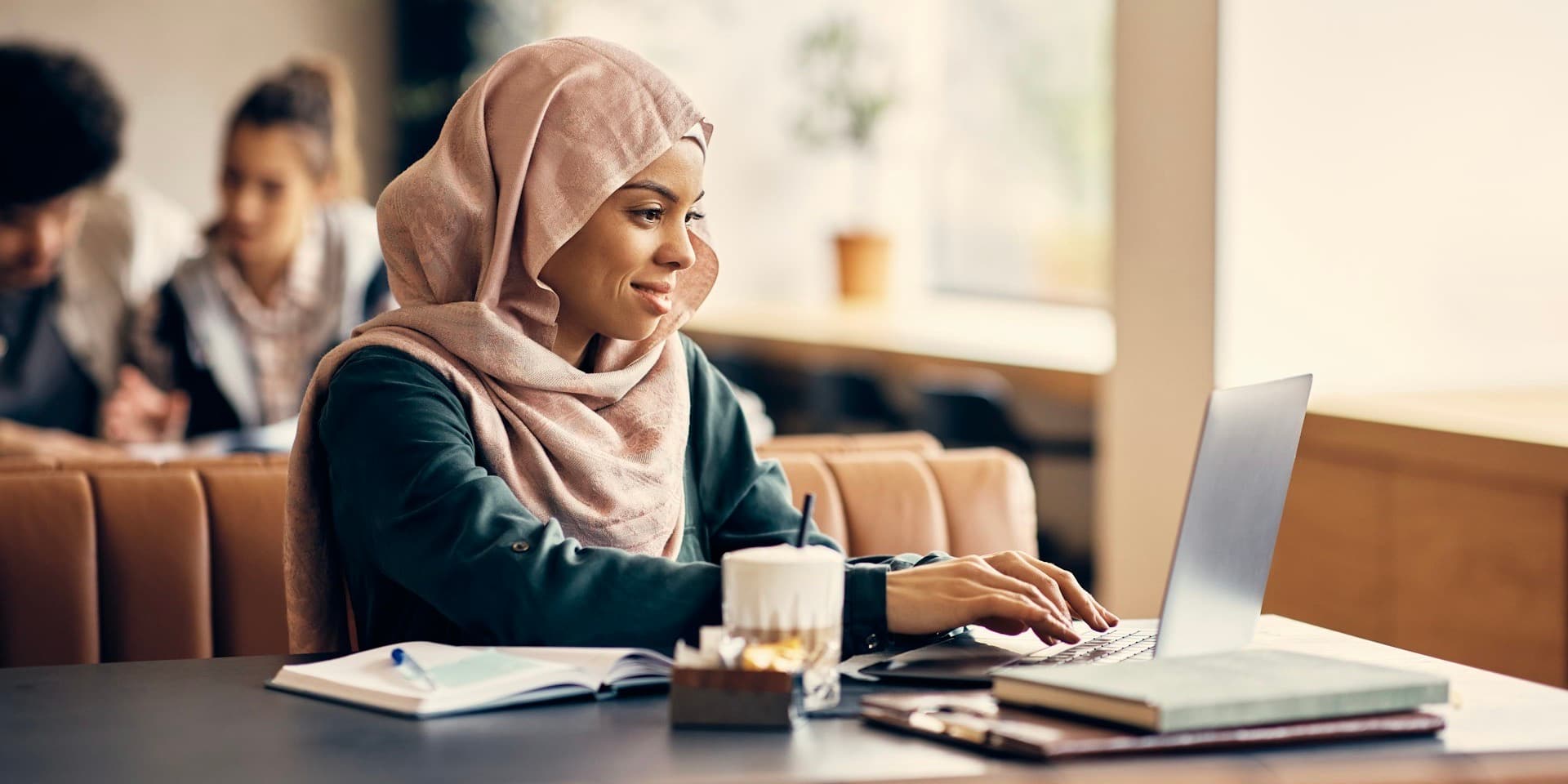 A woman in a pink headscarf and black dress sits in a booth at a restaurant, smiling as she types into a computer, with coffee and books on the table for this Dr. B article on how to use an HSA to save on healthcare costs.