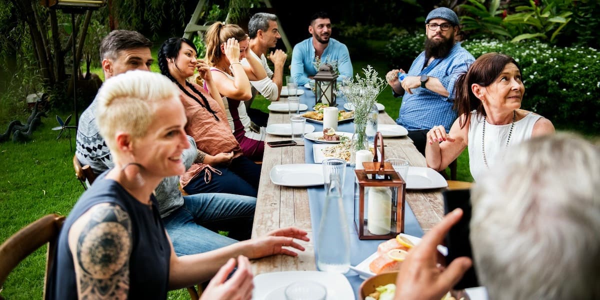 A group of adult friends sit outside at a long wooden able covered in plates, cutlery, flowers and candles, all looking at someone out of frame, for this article about microdosing GLP-1s.