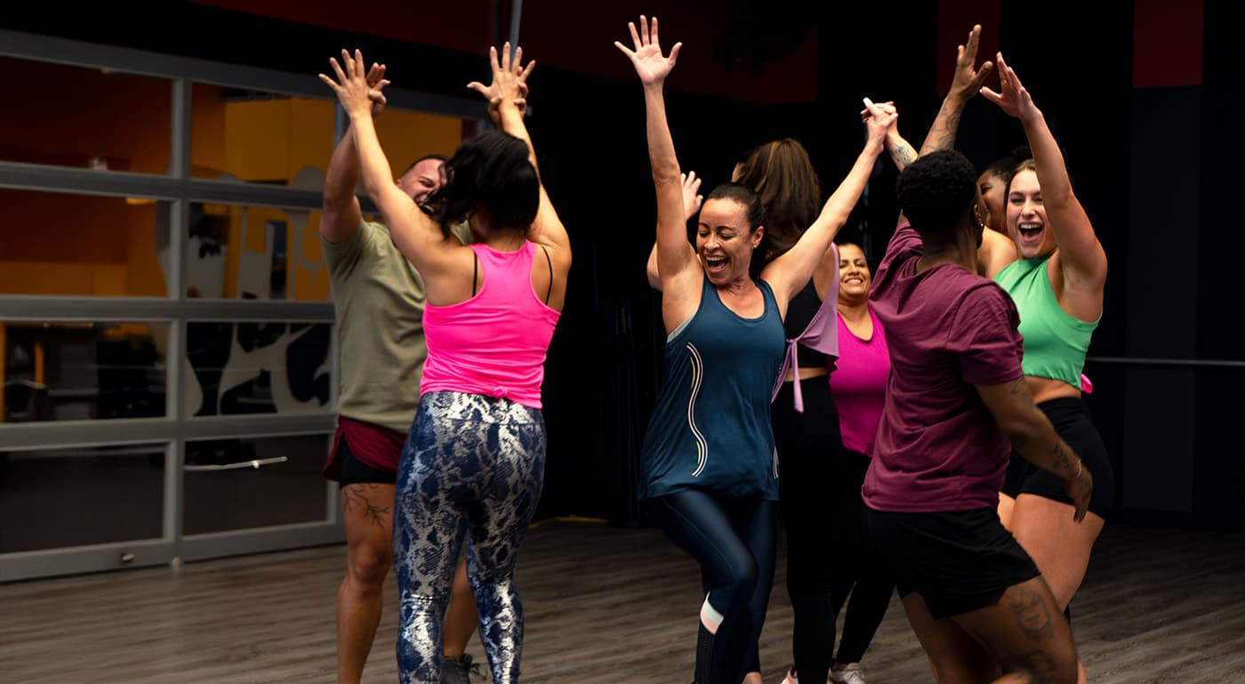 A diverse group of people in athletic wear celebrate together in a fitness studio, smiling with their arms raised in a joyful group moment after a workout.
