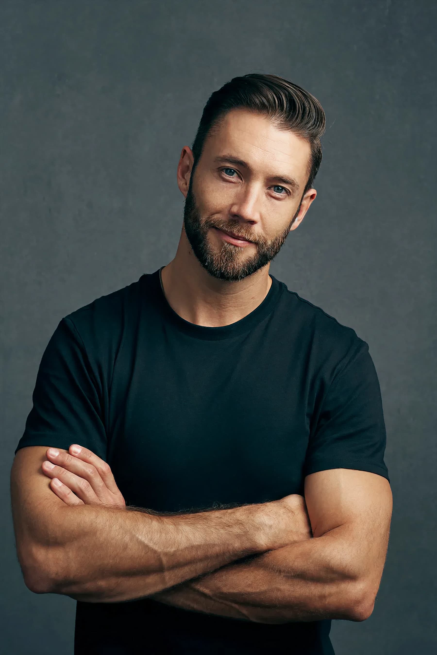 Smiling man with crossed arms in black shirt standing in front of a grey backdrop