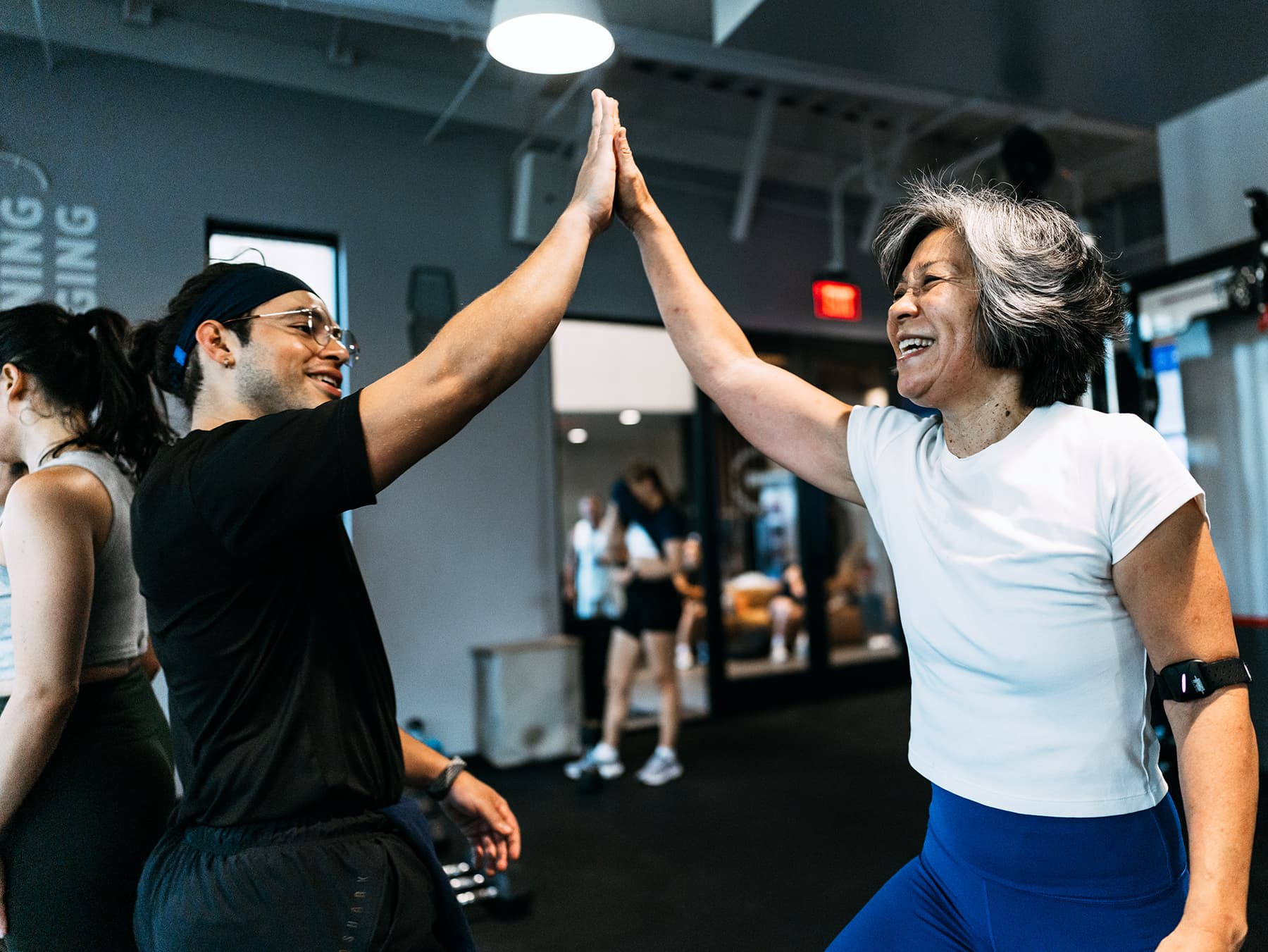 Two people in a gym smile and high-five during a workout, with others exercising in the background.