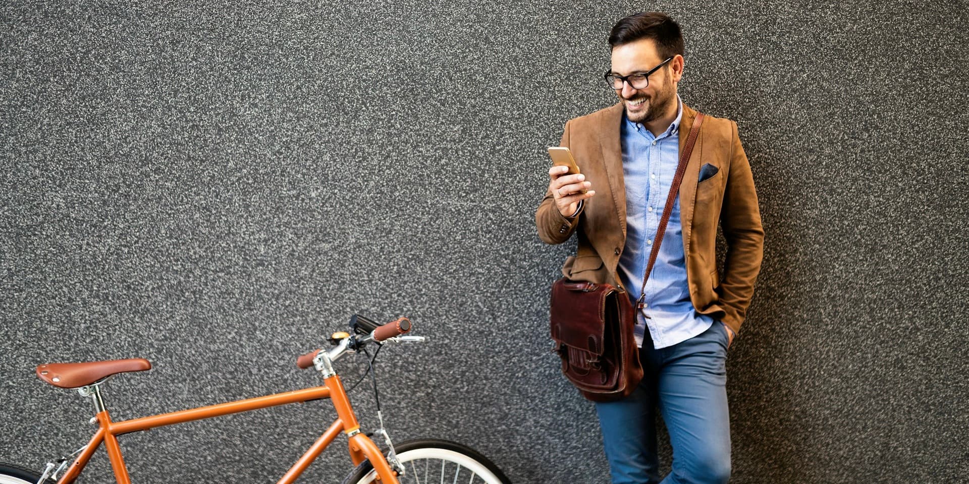 In an article about which is cheaper for online hair loss, a man in casual business attire stands next to a wall, smiling as he looks down at his phone, with a bicycle propped nearby.