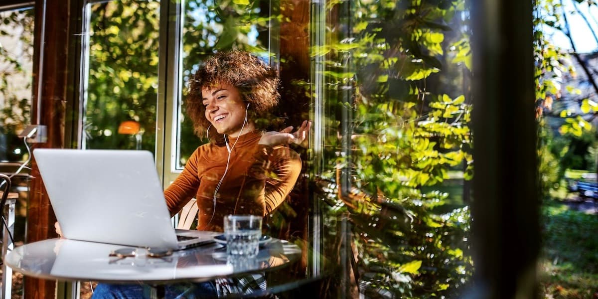 In an article about how health insurance works for Dr. B, a woman sits outside at a cafe table, smiling into a laptop, while bright green trees reflect off of the windows behind her.